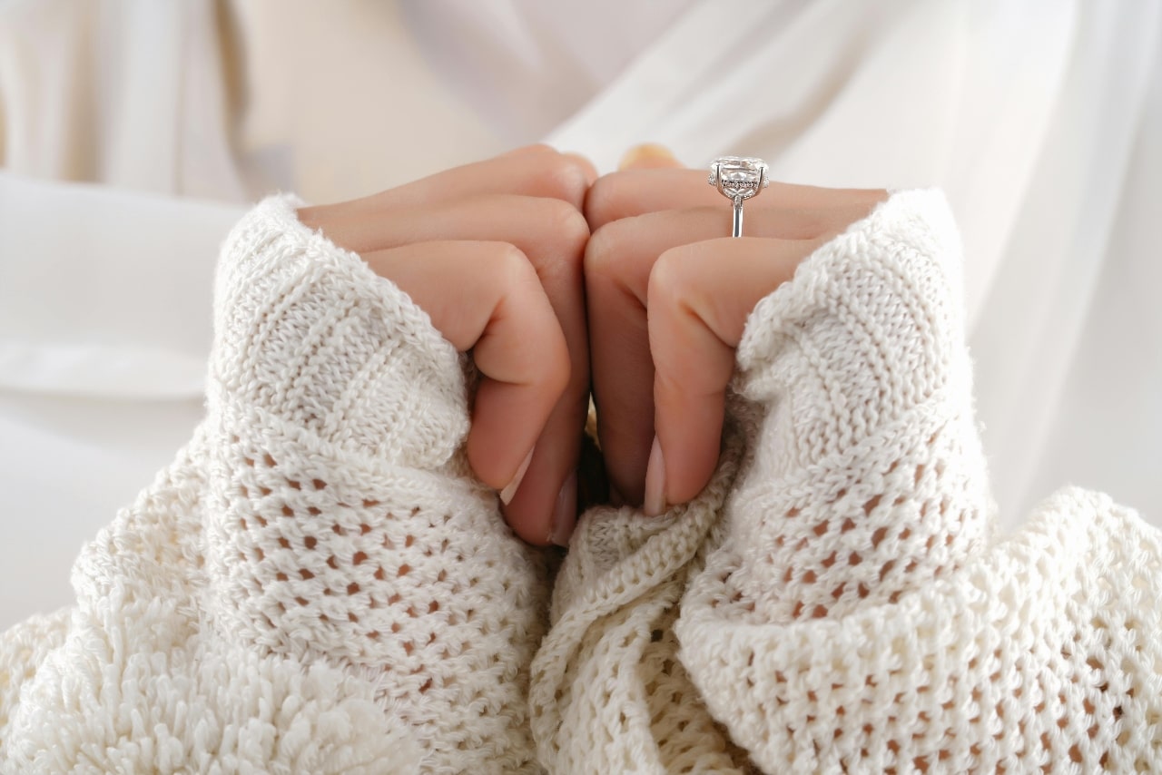 A close-up of a woman’s hands wearing a delicate engagement ring, wrapped in a cozy sweater.