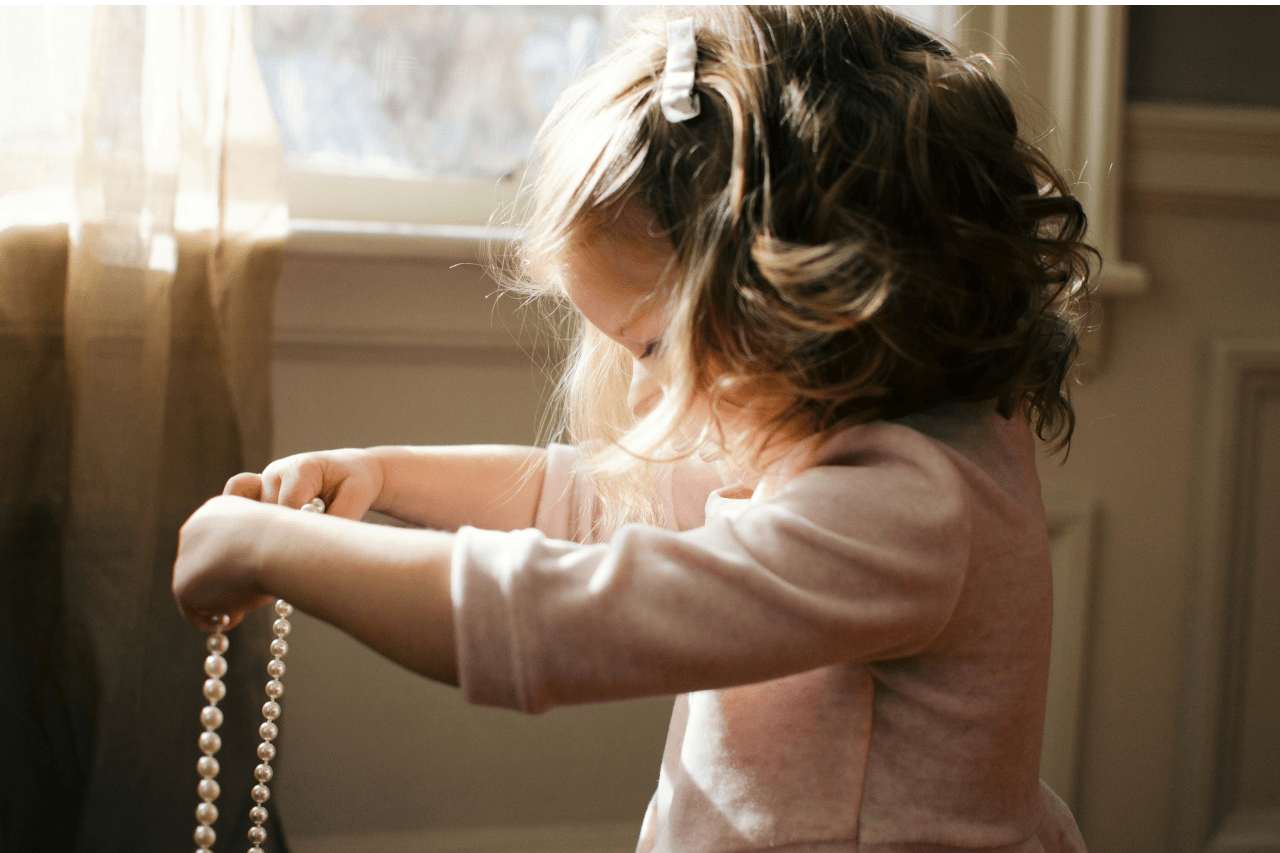 A little girl holding up a string of pearls and standing near a window.