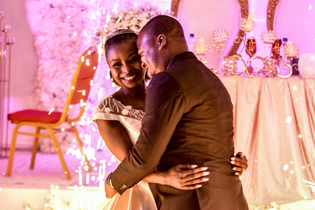 A newlywed couple embracing under romantic pink light at their wedding reception.