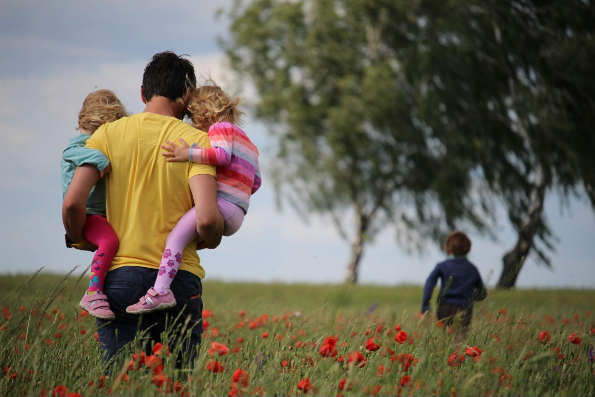 A dad carries two young children through a field while an older boy runs ahead.