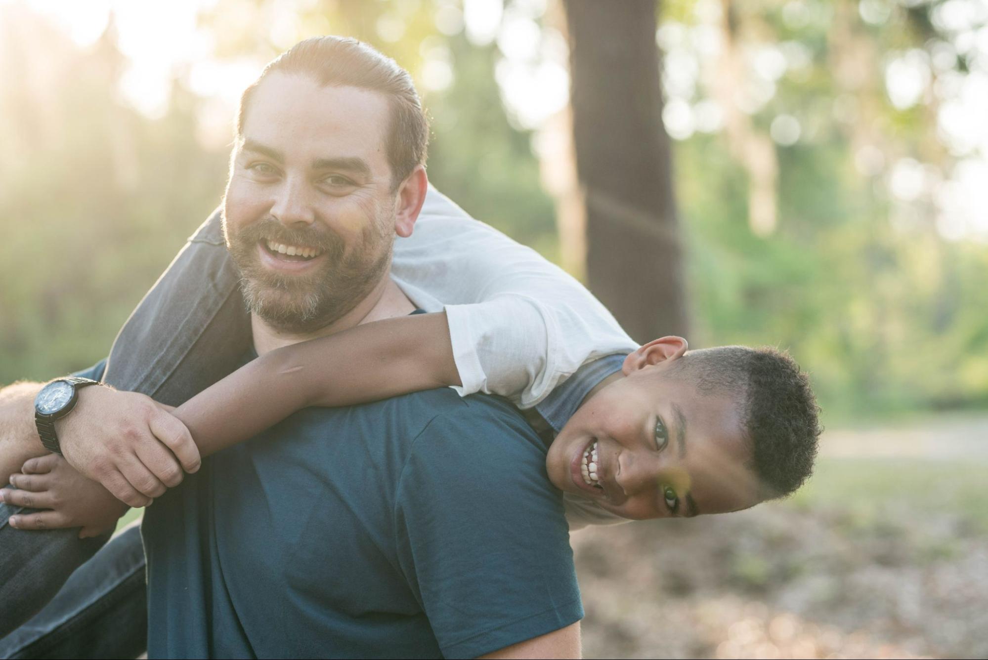 A man hoists his son over his shoulder in a park, both smiling.