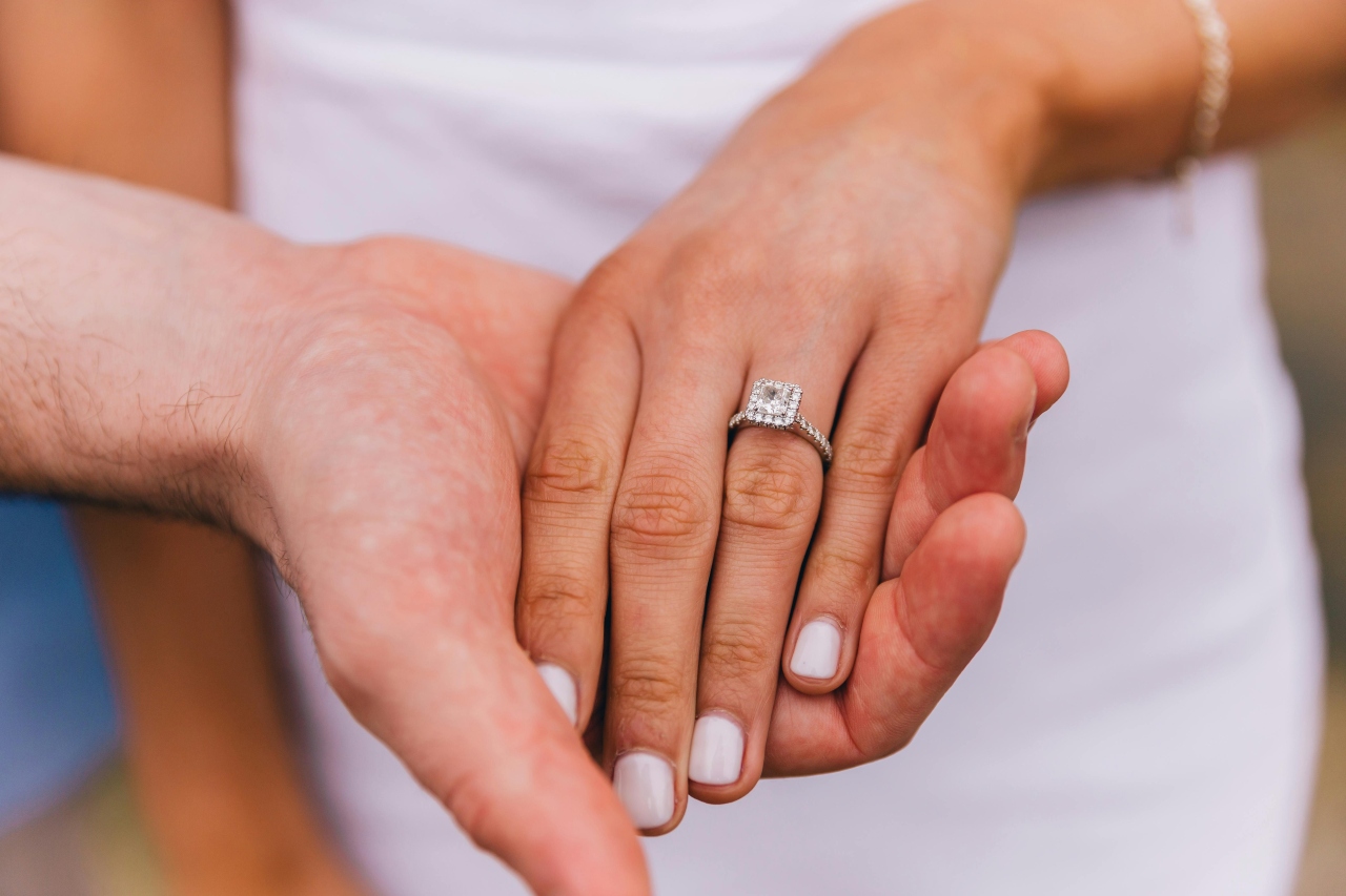 a woman’s hand being held by a man’s and adorned with a princess cut engagement ring