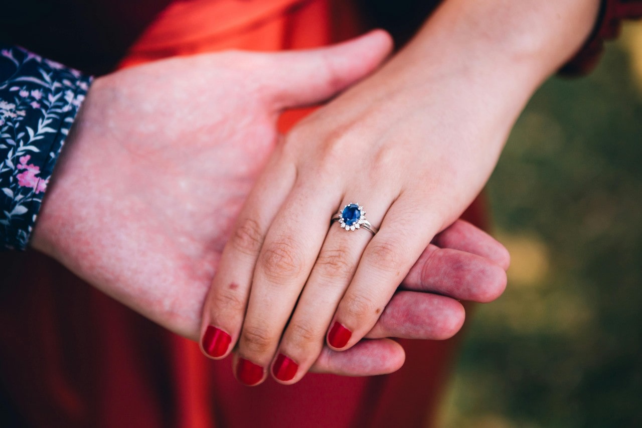 a man’s hand holding a woman’s, which is adorned with a sapphire engagement ring