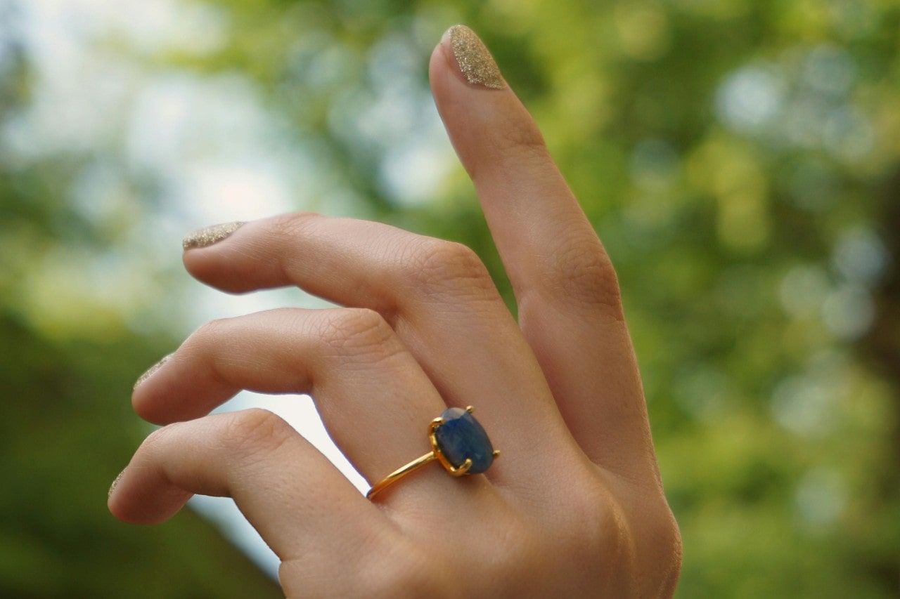 a hand raised in the air, adorned with a solitaire engagement ring featuring a blue opaque stone