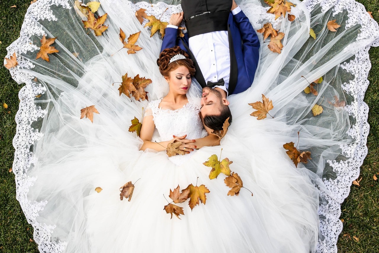 An aerial view of a bride and groom laying on the grass, lightly covered in autumn leaves.