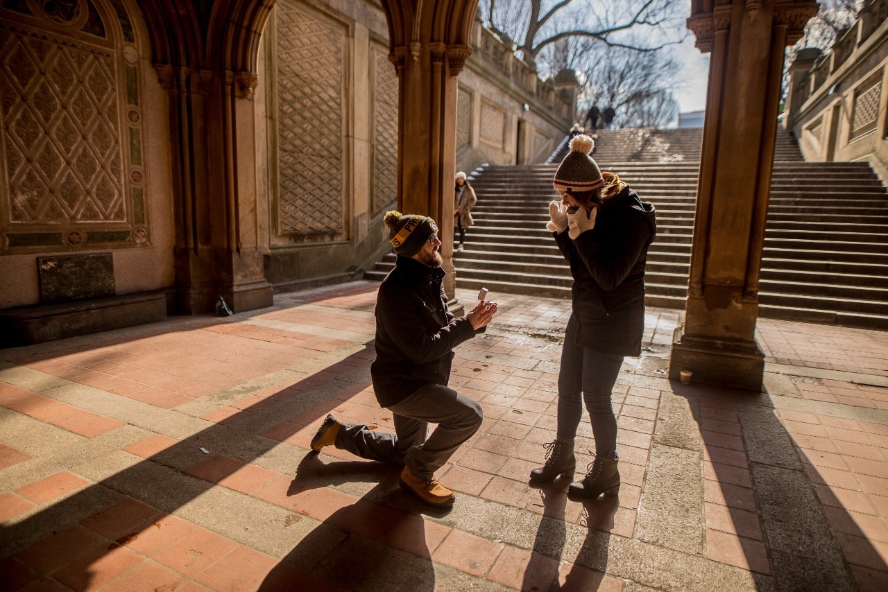 A man proposing to a woman at the end of some stairs in an open plaza on a cold winter morning