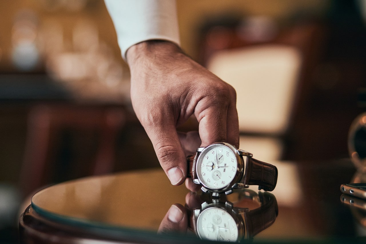 Hand lifting a watch with a white dial and a brown leather strap off of a round table
