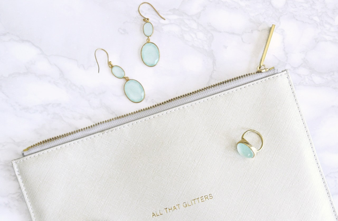 A pair of gold and blue topaz drop earrings sit outside a white leather pouch with gold lettering and a blue topaz bezel fashion ring