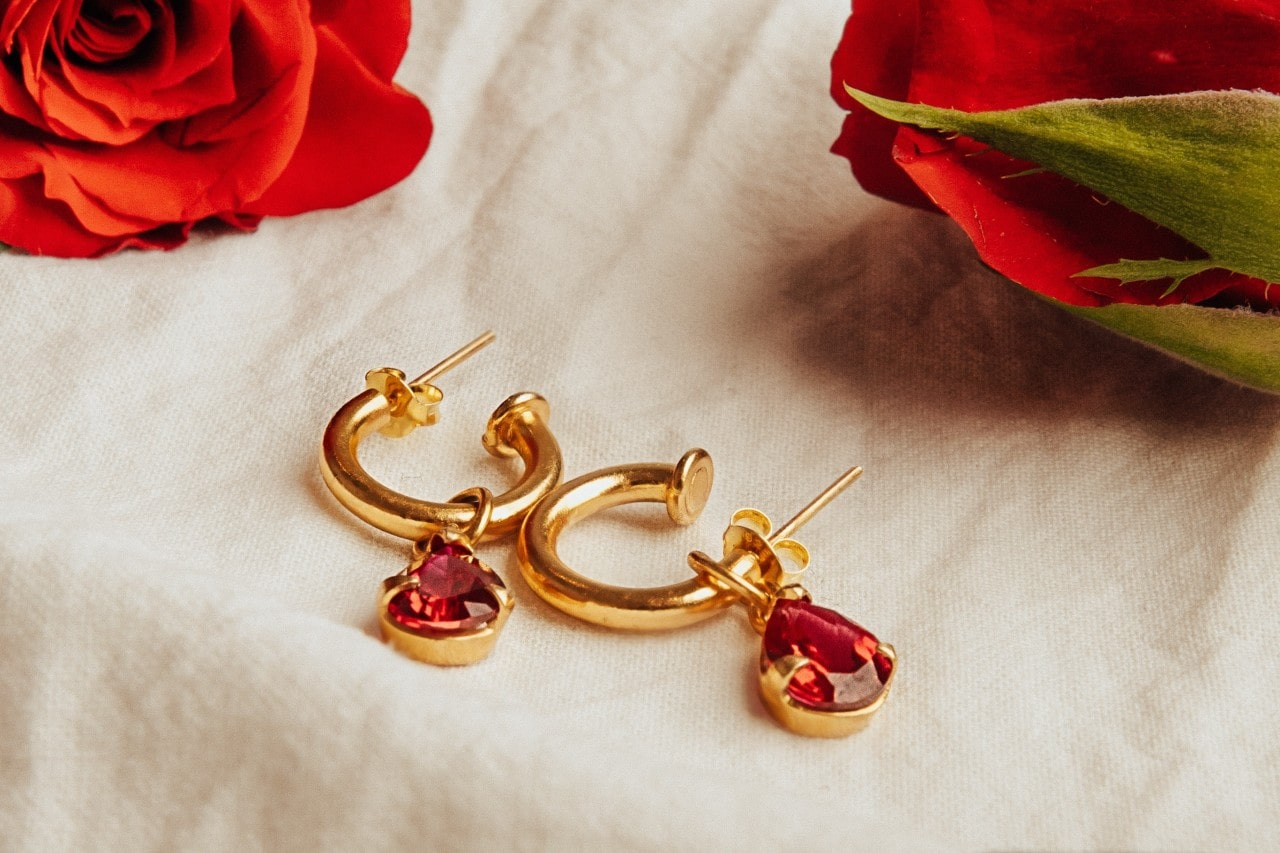 A pair of yellow gold huggie earrings with a ruby gemstone accent sits on a cream bedsheet with roses