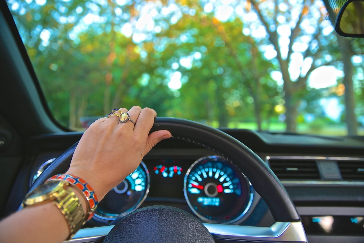 A woman wearing a yellow diamond fashion ring, a colorful enamel bangle, and a yellow gold timepiece drives her SUV down a peaceful country road