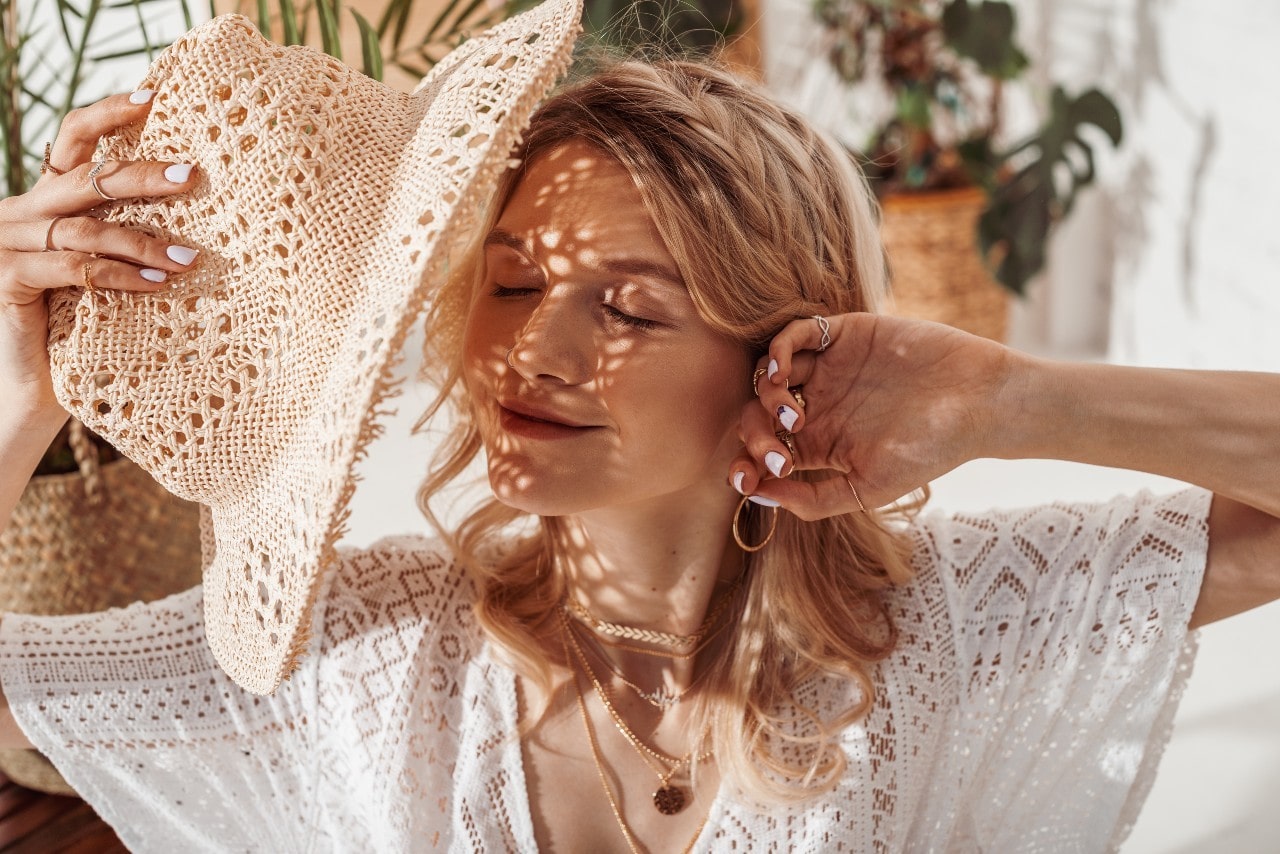 A woman wearing jewelry stretches and shields her face from the summer sun with a straw hat