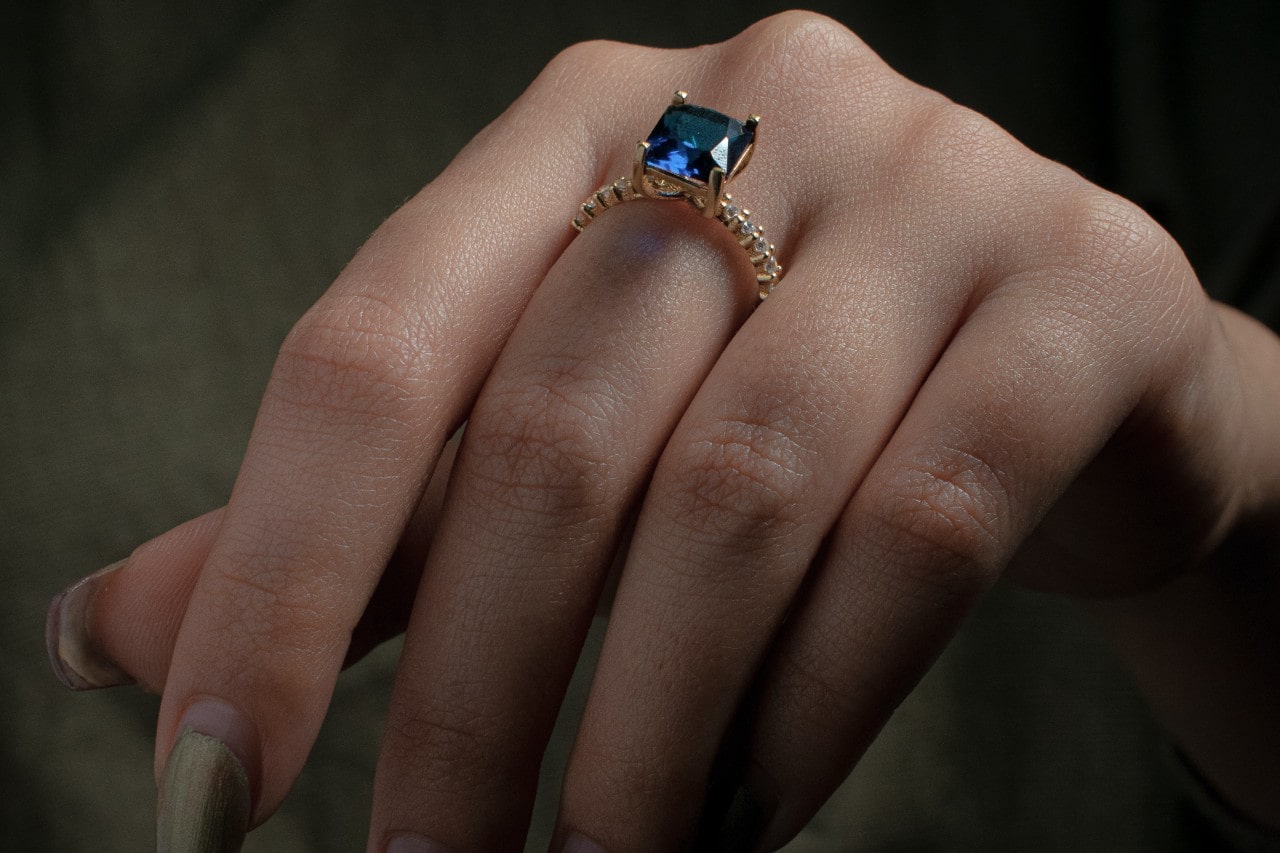 close up image of a hand wearing a yellow gold ring featuring a princess cut sapphire