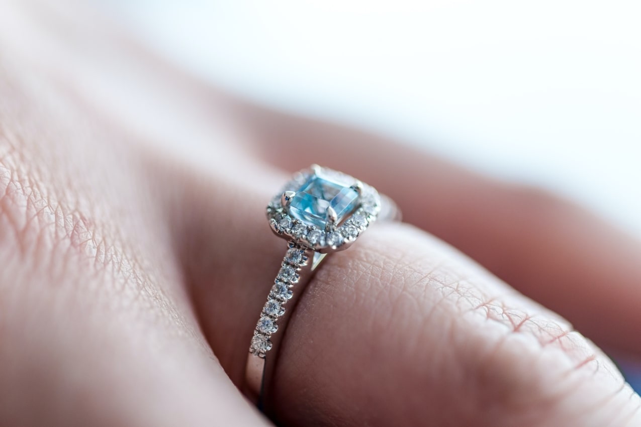 close up image of a hand wearing a silver engagement ring featuring a blue, princess cut center stone