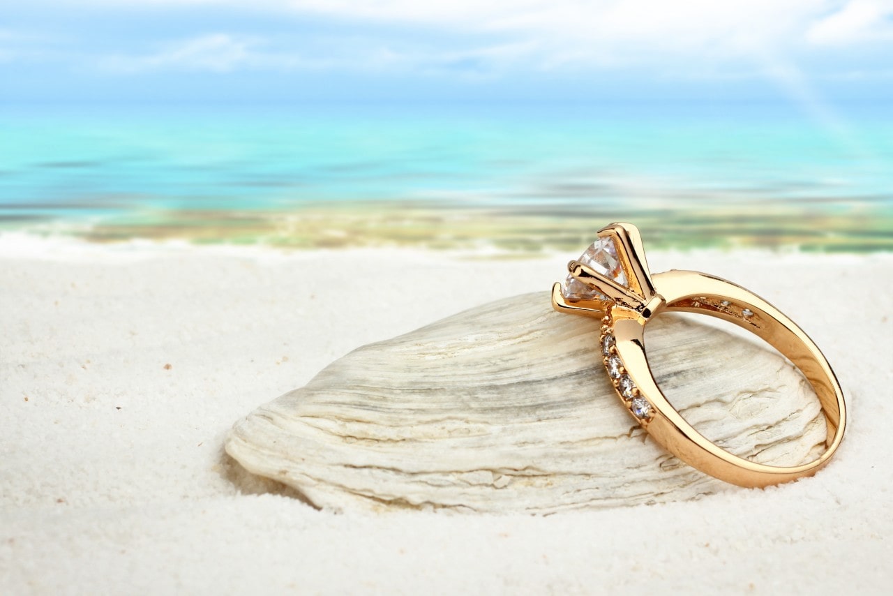 A stunning yellow gold engagement ring resting on a clamshell at the beach.