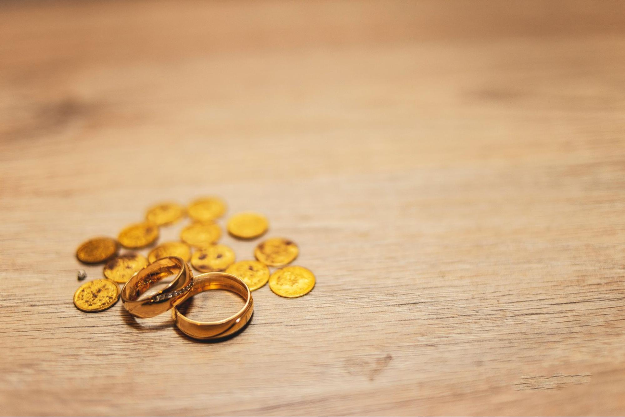 Two gold rings sit by gold coins on a wooden table.