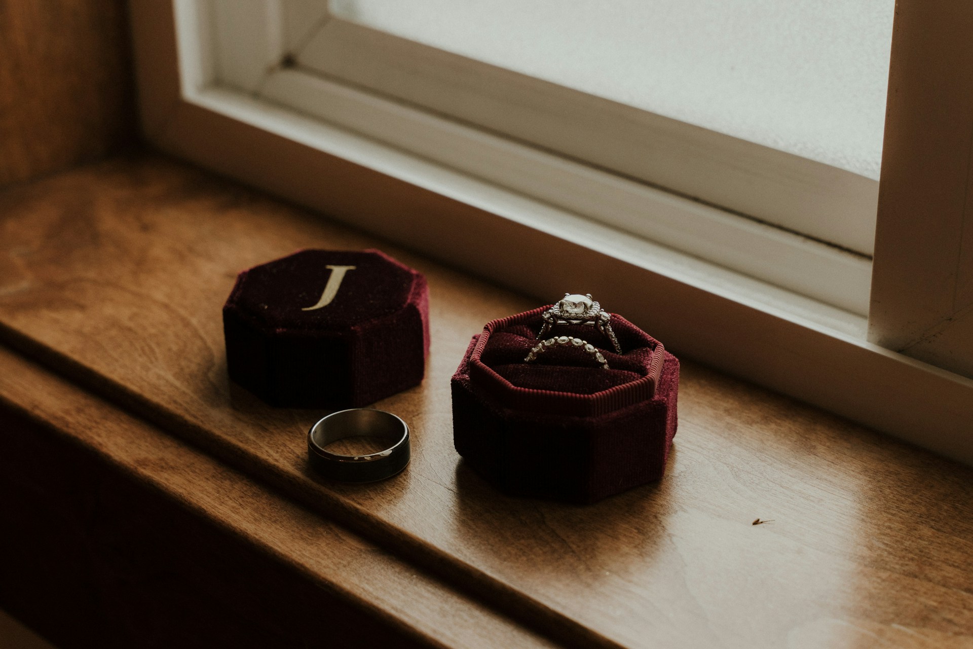 A close-up of a matching white gold engagement ring and wedding band in a red velvet box, resting on a wooden windowsill.